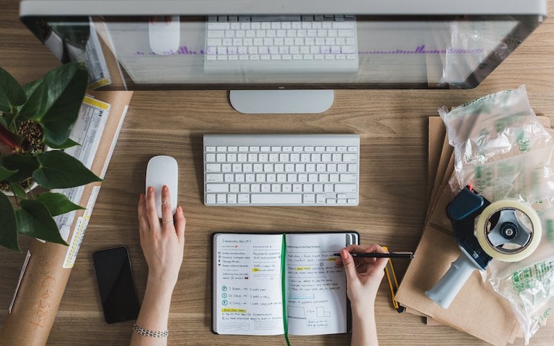 A person writes in a notebook while working from a desktop computer