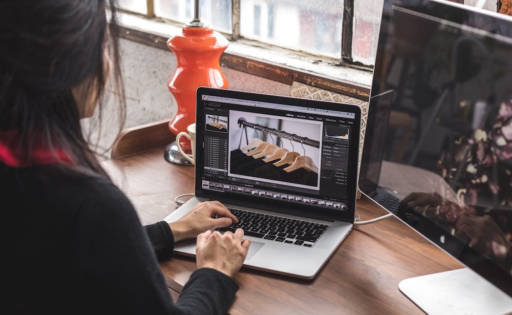 A woman sits in front of a bright window while editing images on her laptop.