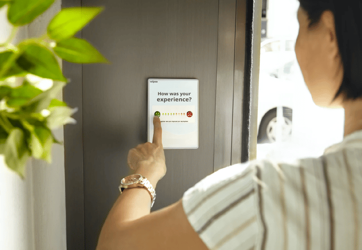 A women leaves feedback on a wall-mounted tablet which prompts 'how was your experience'.