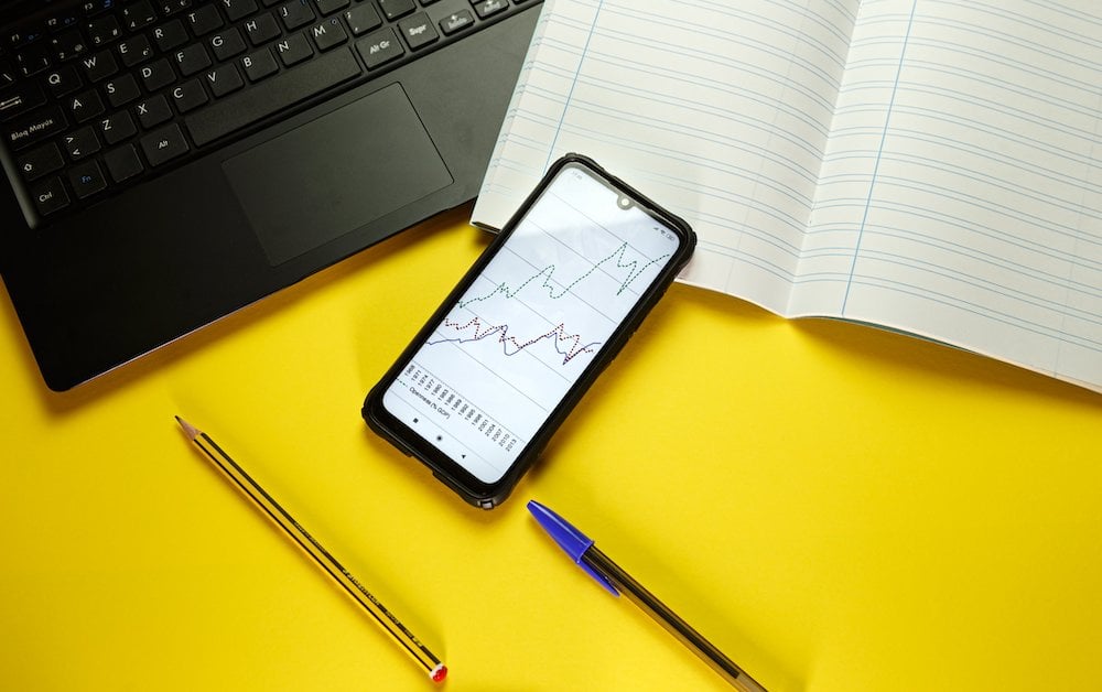 Flatlay of a laptop and open notebook with a cellphone showing graphs and a pen and pencil laying on a bright yellow background