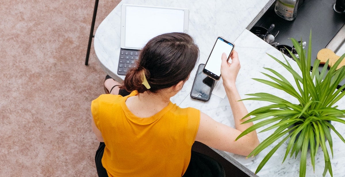 A young woman uses a cellphone while sat at a table in front of a tablet.