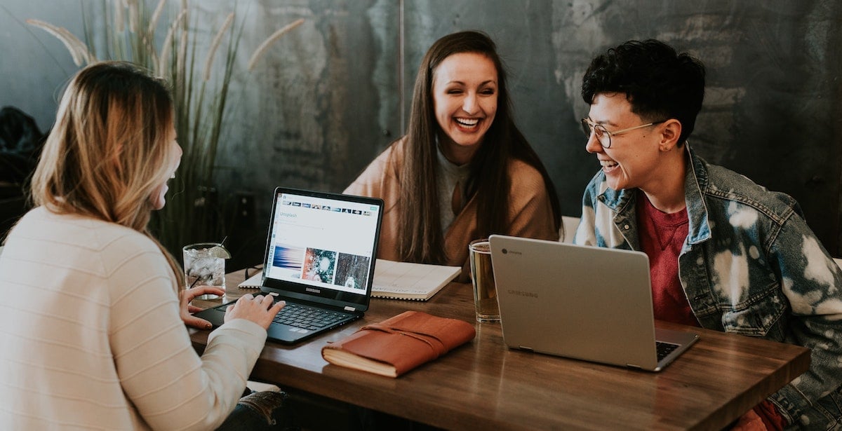 3 women laugh and socialise while sat at a table working from laptops.