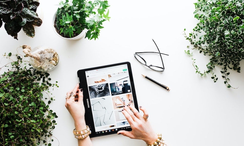 Birds-eye shot of feminine hands holding a tablet and scrolling through images. House plants sit on the table around the hands.